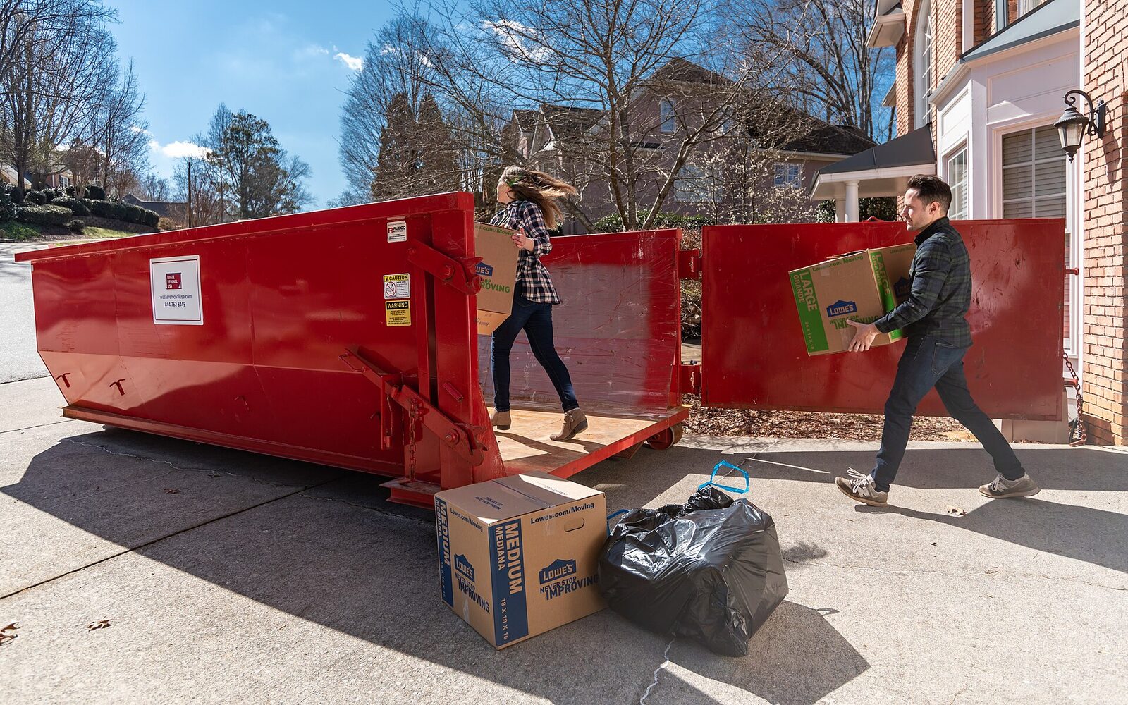 Couple carrying boxes into a red rear-door roll-off dumpster on a suburban driveway