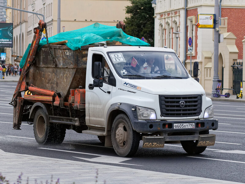 White GAZon skip-loader truck carrying a covered skip bin on the road