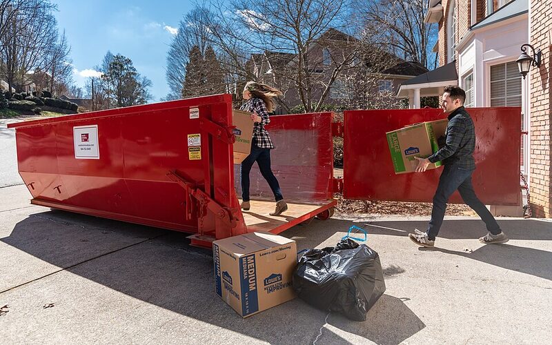 Couple carrying boxes into a red rear-door roll-off dumpster on a suburban driveway